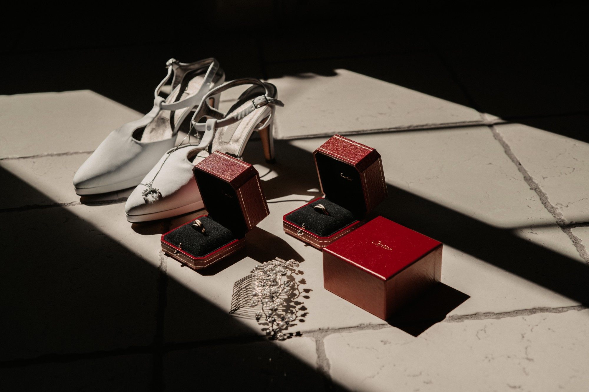 Chaussures de la mariée, alliances dans leurs écrins rouges et peigne de perles dans un rayon de lumière