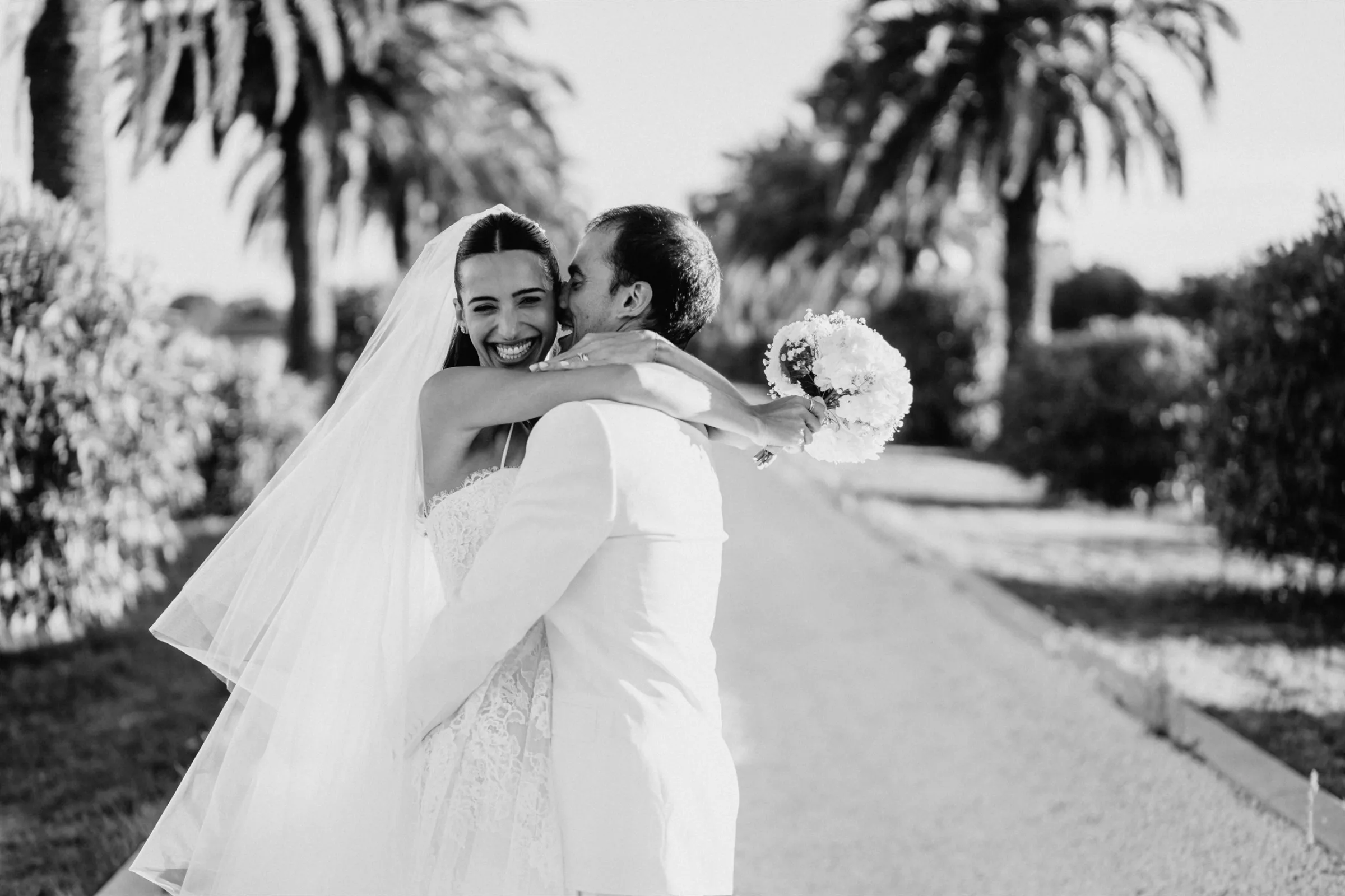 Mariée éclatant de rire dans les bras de son marié, allée de palmiers dans le Var, photo noir et blanc
