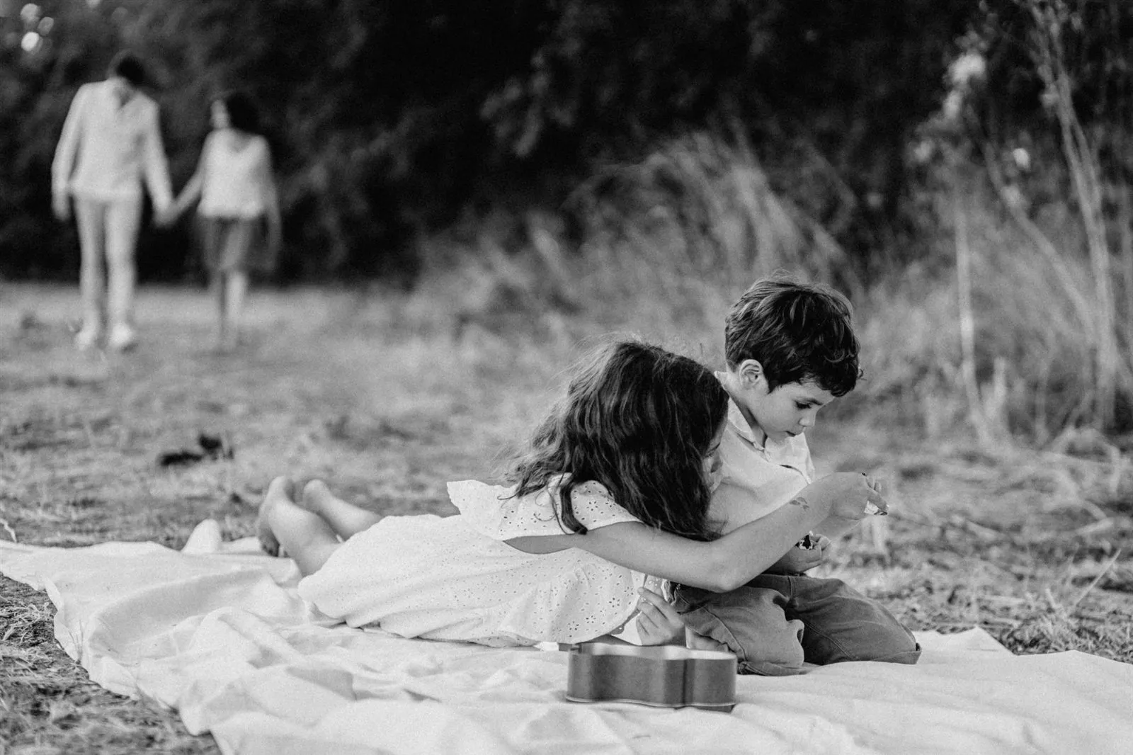Séance photo famille : frère et sœur sur une couverture pendant que les parents s’éloignent, photo noir et blanc